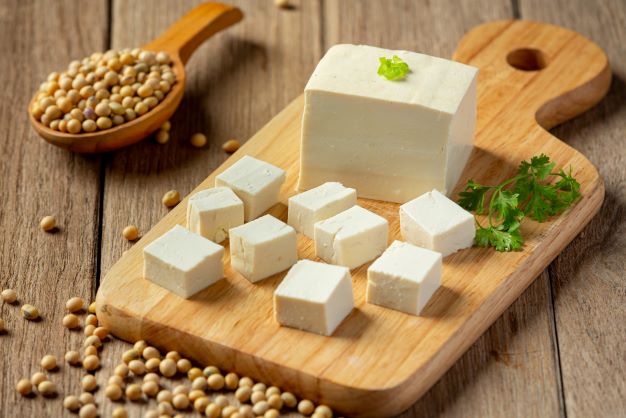 tofu je na drevenej podložke nakrájané na kocky, na vrchu je petržlenová vňať, vedľa je drevená lyžica so sójovými bôbmi. Všetko je to servírované na drevenom stole