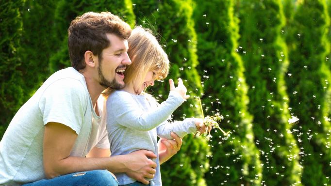 Otec s malou dcérou sa smejú a dcéera sa hrá s odvkitnutou púpavou, za nimi sú zelené stromy.