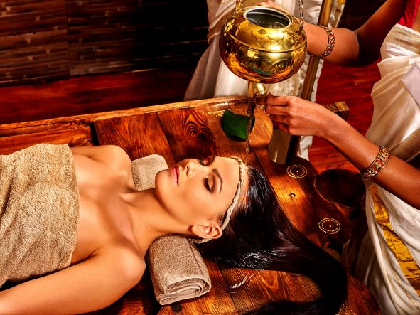 krásna žena ležiaca na drevenom stole realaxuje a na hlavu jej tečie teplý bylinkový olej medenej nádoby
