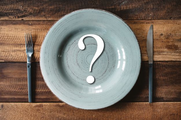 tanier šedej farby je položený na drevenom stole s vidličkou a nožíkom, v strede taniera je veľký biely otáznik.
