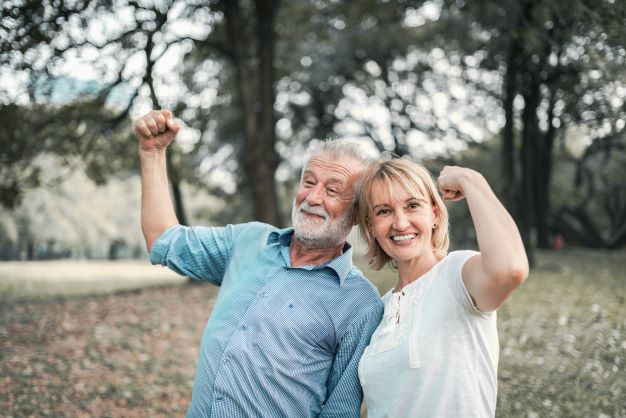 starší muž v modrej košeli stojí so ženou v bielom tričku v prírode, ukazujú, že sú silní a zdraví napnutím bicepsu a usmievajú sa.