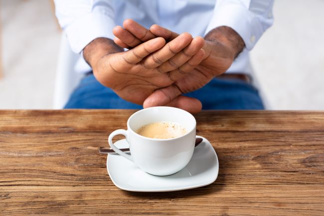 Biela šálka s kávou na drevenom stole, nad ňou sú prekrížené mužské ruky ako náznak odmietnutia šálky s kávou.