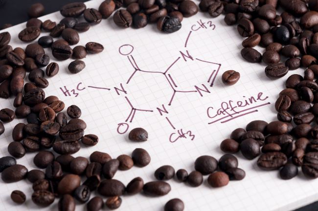 Na kockovanom papieri je napísaný chemický vzorec kofeínu, okolo sú hnedé kávové zrná.