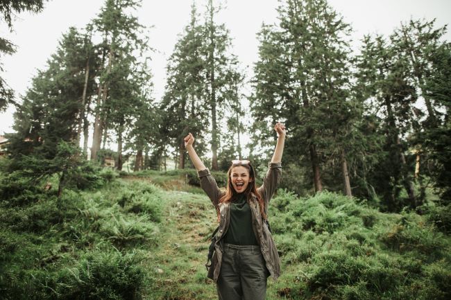 Mladá žena stojí na kopci v lese, ruky má zdvihnuté hore ako náznak slobody.