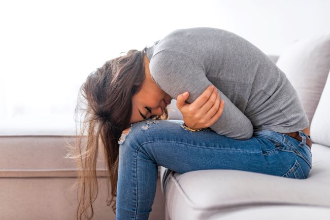 Žena v sivom tričku a modrých rifliach sedí v bolesti prehnutá na bielom gauči a rukami si drží oblasť žalúdka.