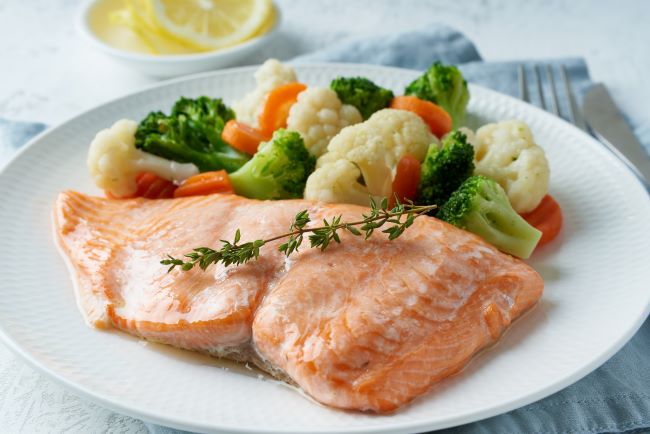 FOODMAP jedlo. Na bielom tanieri je naservírovaná dusená zelenina na pare, brokolica, karfiól a mrkva. Steak z lososa ružovej farby je ozdobený vetvičkou tymiánu. 