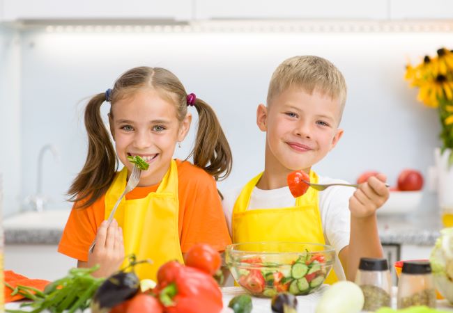 Blonďavý chlapček a hnedovlasé dievčatko s dvoma copíkmi sedia za stolom, kde je veľa zeleniny, obaja sa usmievajú a jedia zeleninový šalát.