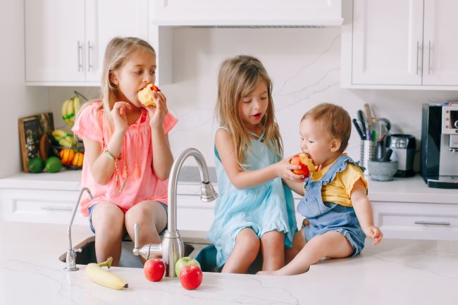 Tri malé deti sedia na kuchynskej linke. Blondína hryzie jablko, hnedovláska kŕmi najmladšie dieťa broskyňou.