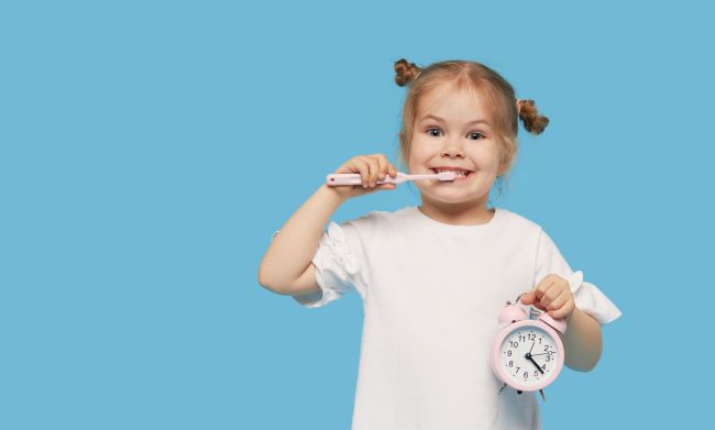 Malé blonďavé dievčatko v bielom tričku drží v jednej ruke ružové budíkové hodiny a v druhej má zubnú kefku, umýva si zuby a usmieva sa.