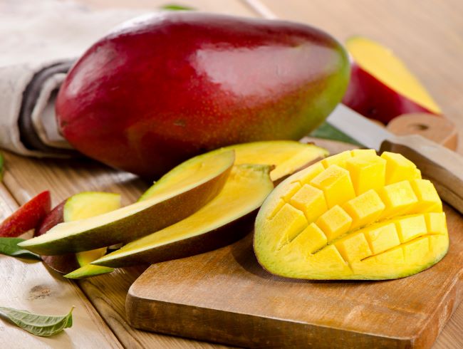 Dozreté žlé mango je na drevenom podnose nakrájané na kocky, vedľa je položené celé mango v červeno - zelenej šupke.
