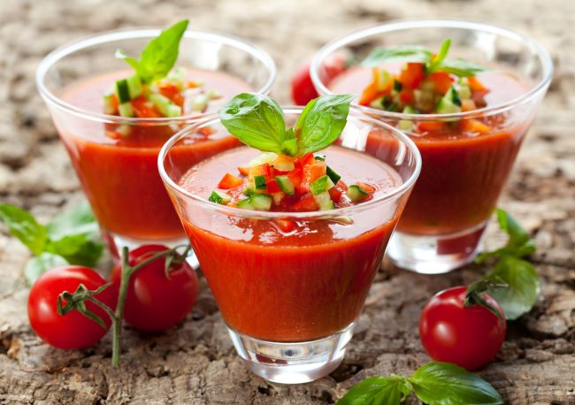 Gazpacho nápoj, z paradajok je naservírovaný v sklenených pohároch s čerstvou bazalkou na drevenom stole, okolo sú poukladané paradajky.